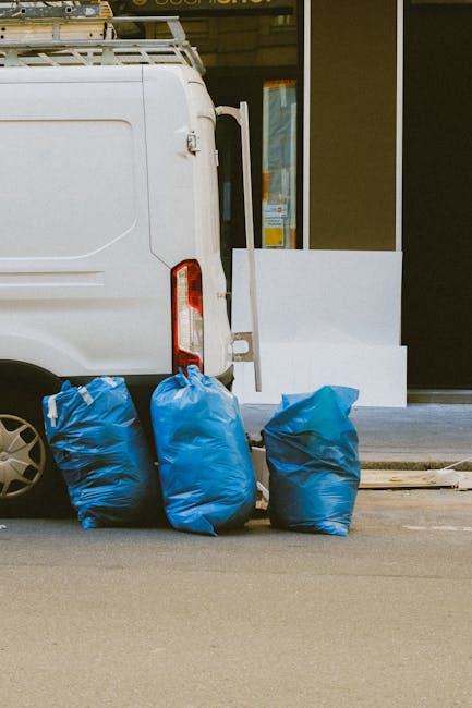 In the image, three large blue rubbish bags made of plastic are positioned on the pavement next to the rear of a white commercial van, which is parked possibly on a residential or commercial street. T