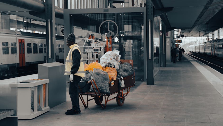 A person wearing a grey knit cap, high-visibility vest, and dark clothing, standing beside a red metal trolley filled with various black, grey, and orange plastic bags and household waste, is position