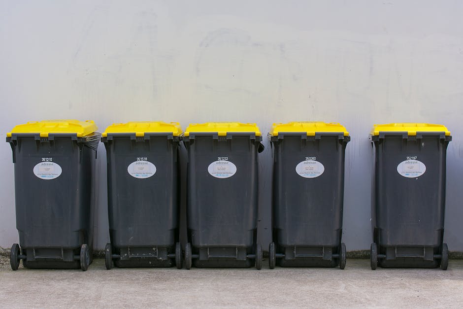 A person wearing a grey knit cap, high-visibility vest, and dark clothing, standing beside a red metal trolley filled with various black, grey, and orange plastic bags and household waste, is position